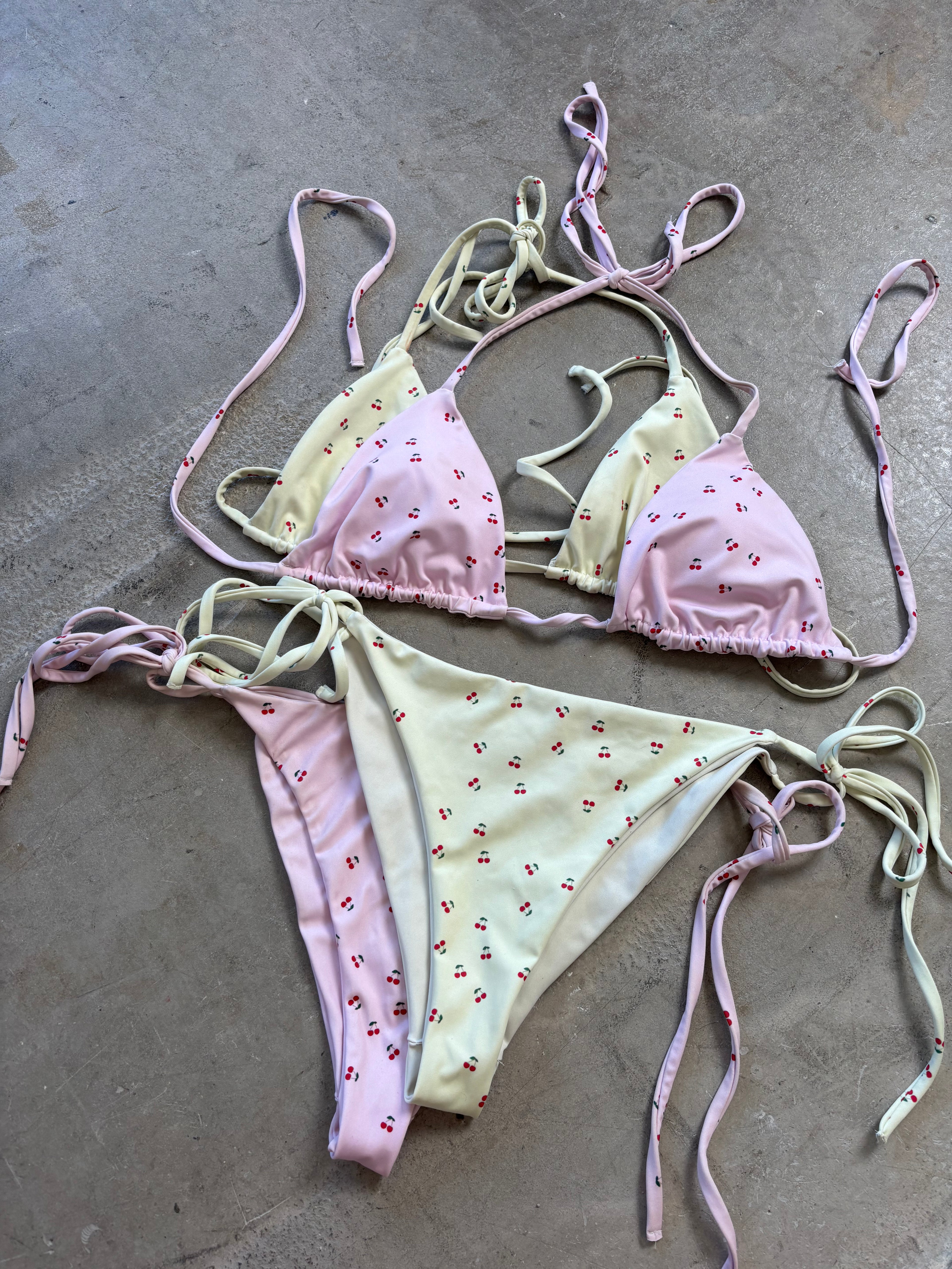 A yellow bikini bottom called the Cherry Orchard, made from recycled polyester with UPF 50+ sun protection. The bottoms feature a refreshing cream base with a vibrant cherry pattern and are available in sizes 2XS to 6XL. They are designed with soft, stret